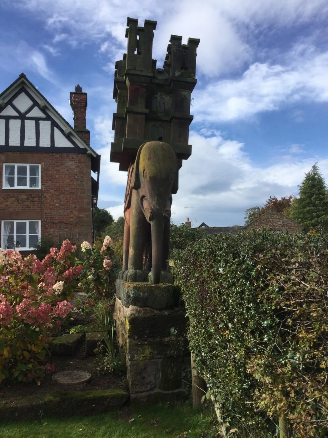 The sandstone elephant before the rescue, carrying a carved stone castle on its back and standing on a worn sandstone base, between a red-brick house and a holly hedge